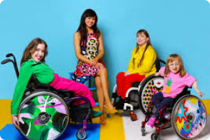 Four women, 3 in wheelchairs showing the multi coloured designs of available wheelchair covers